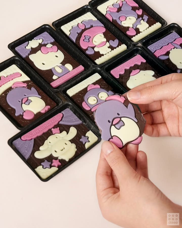 산리오 캐릭터즈가 그려진 초콜릿 8개가 테이블 위에 놓여 있고, 우하단 사람 손이 캐릭터 선을 따라 초콜릿을 부수고 있는 모습