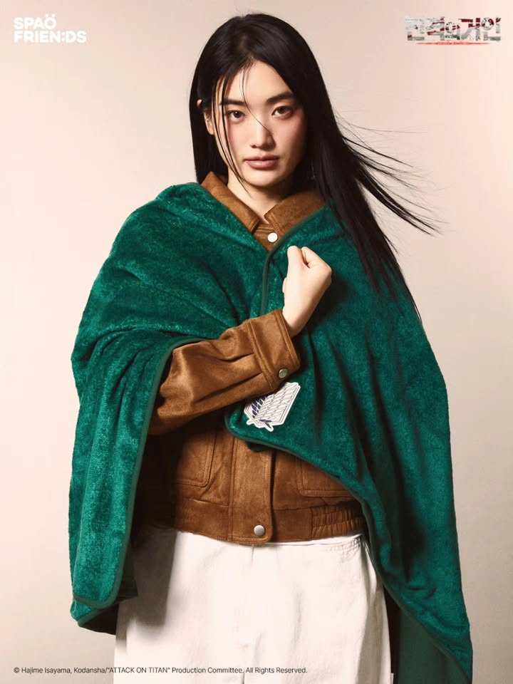 상단 'SPAO FRIENDS 진격의 거인' 텍스트. 중앙에 모델이 갈색 자켓과 초록색 진격의 거인 후드 담요를 두르고 심장 쪽에 주먹을 갖다댄 채 서 있는 모습.