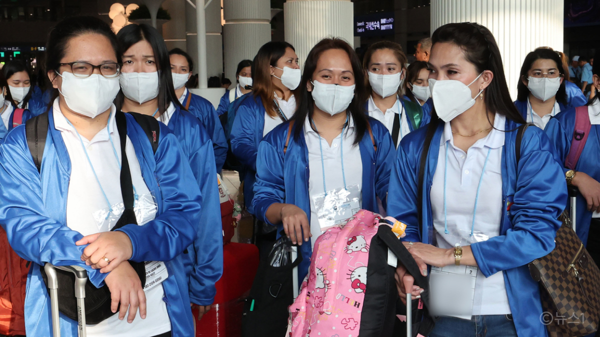 필리핀 여성들이 '외국인 가사관리사 제도'로 인천공항에 입국하는 모습이에요. 여성들은 흰 티셔츠와 파란색 자켓을 입고 캐리어를 끌며 걷고 있어요.