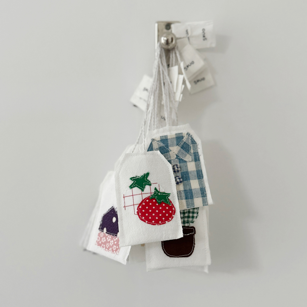 흰 바탕에 북-티 제품이 여러 개 걸려 있는 모습이에요. 책갈피 위에는 토마토, 셔츠 등의 모양이 박음질돼 있어요.